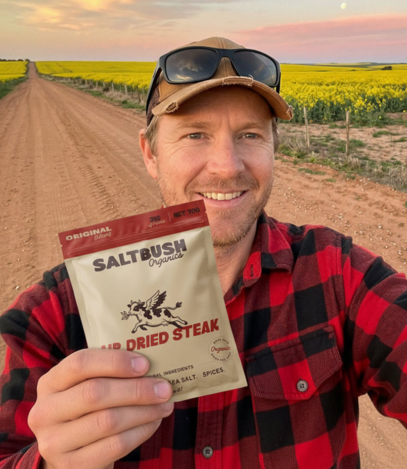 Man holding a packet of Saltbush Organics dried steak in front of a yellow field and dirt road.