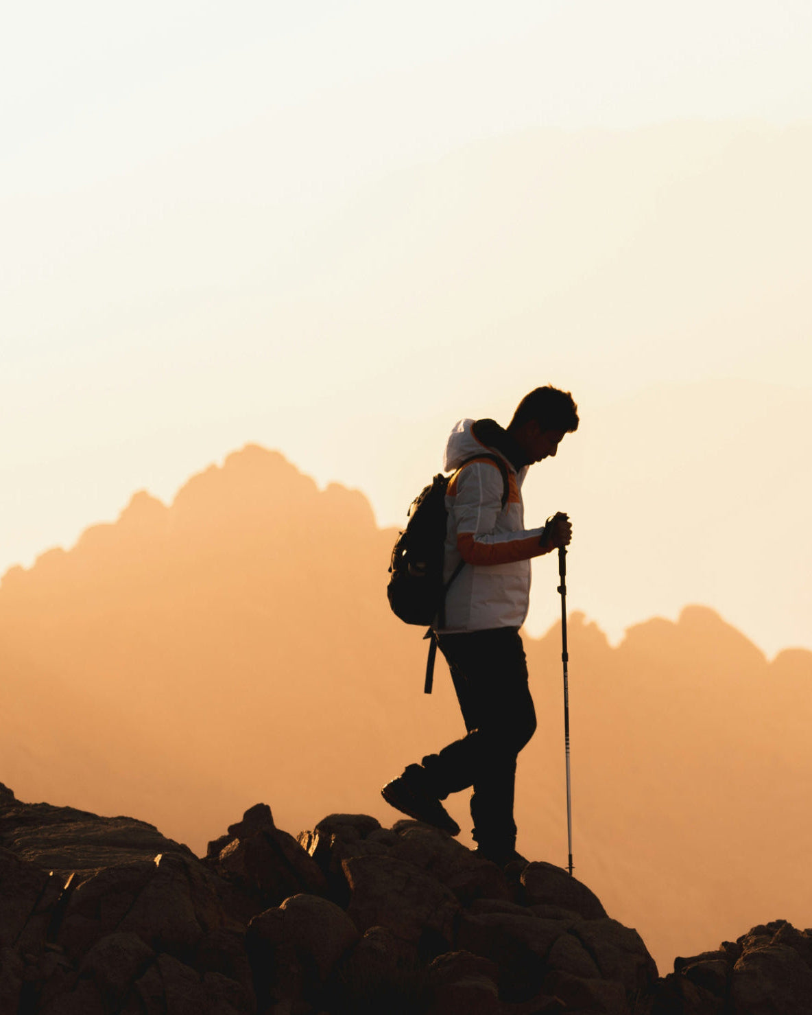 Silhouette of a hiker with a backpack and walking stick against a mountainous landscape during sunset.