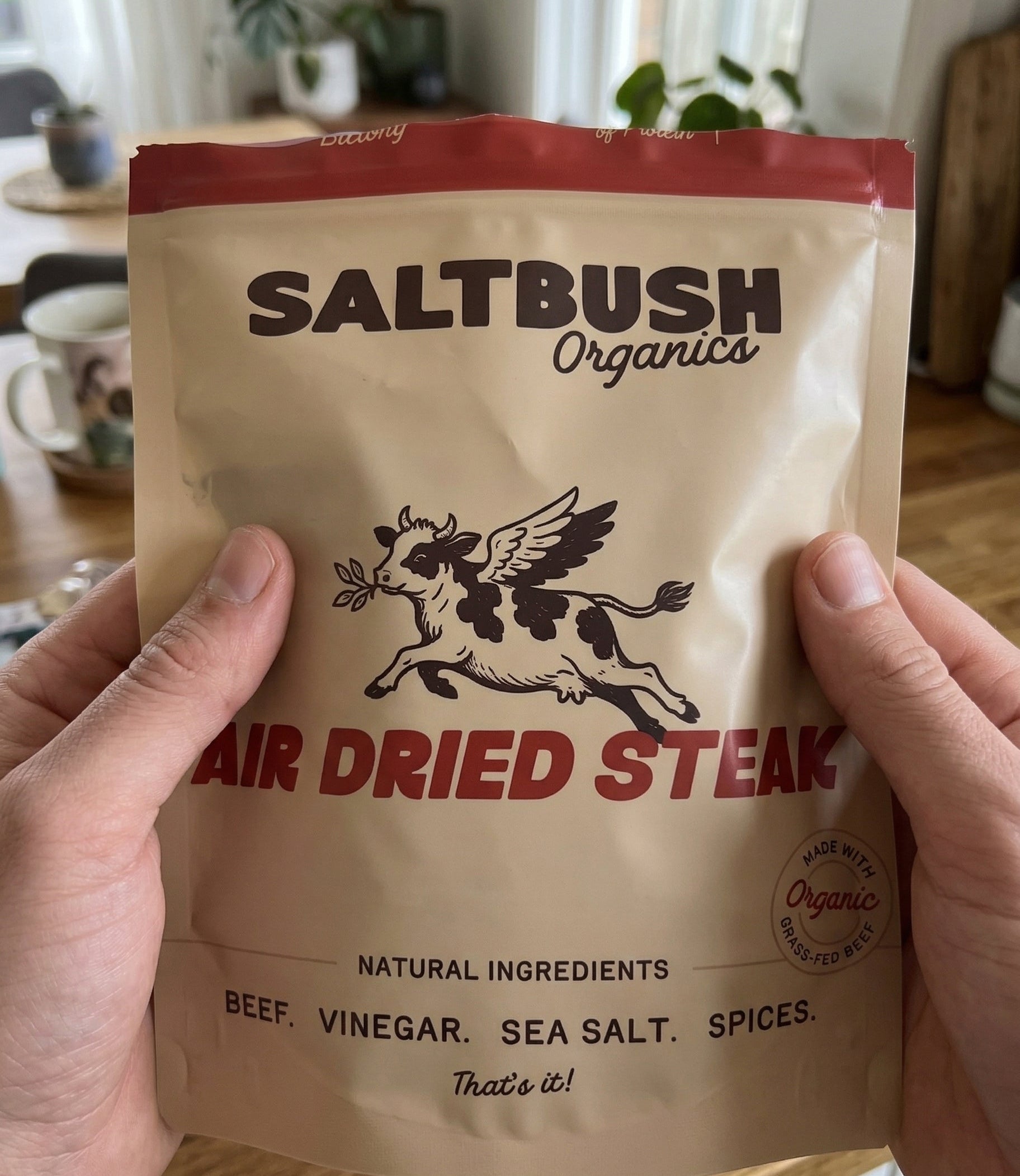 Saltbush Organics Biltong air dried steak package held by a person in a kitchen setting.